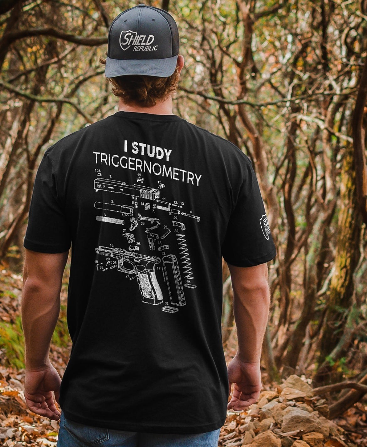 Person wearing a black t-shirt with gun diagram and text, standing in a forest.