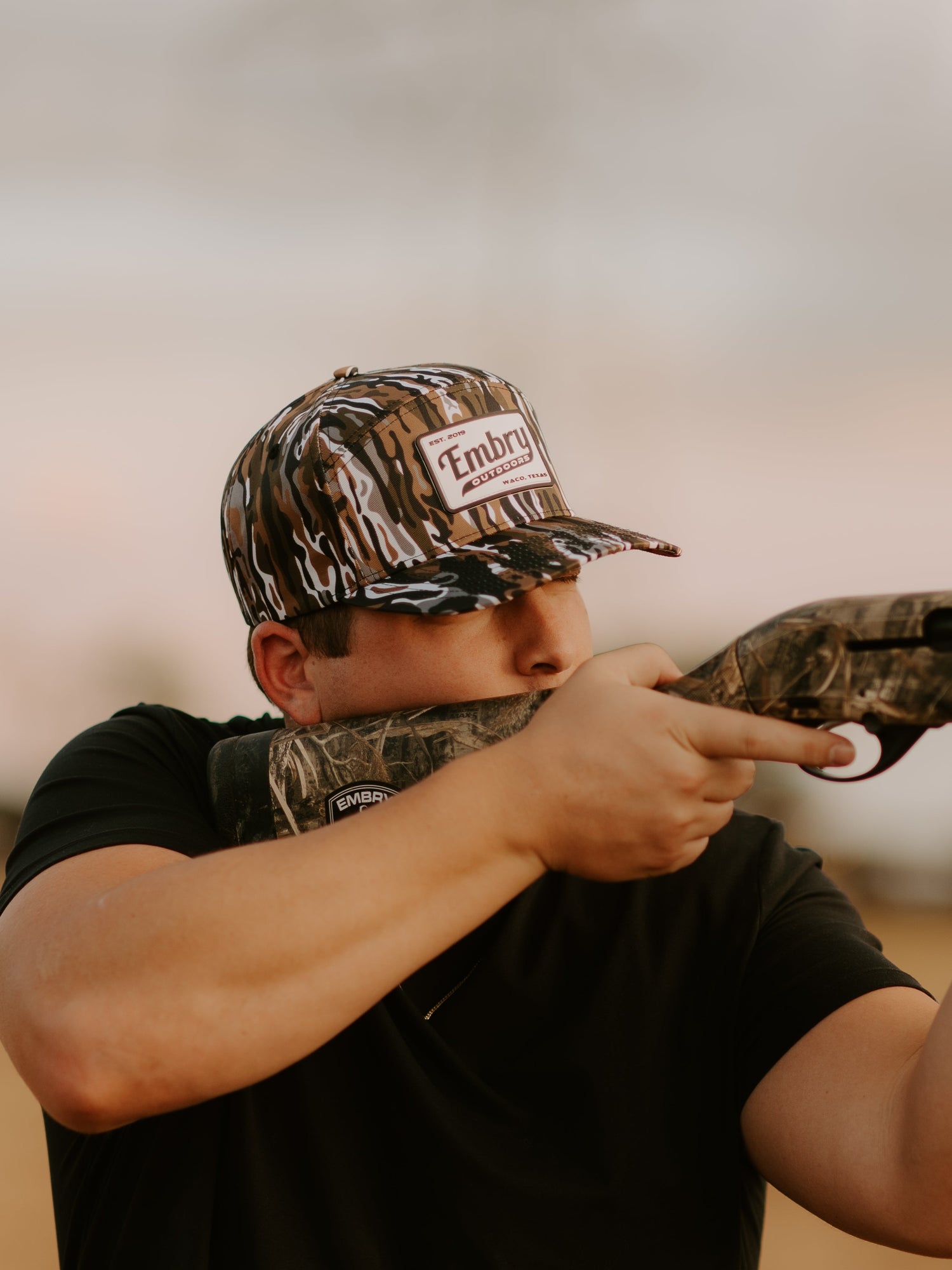 Man wearing camouflage hats aiming with a rifle outdoors, showcasing durable camo headwear for outdoor activities and hunting.