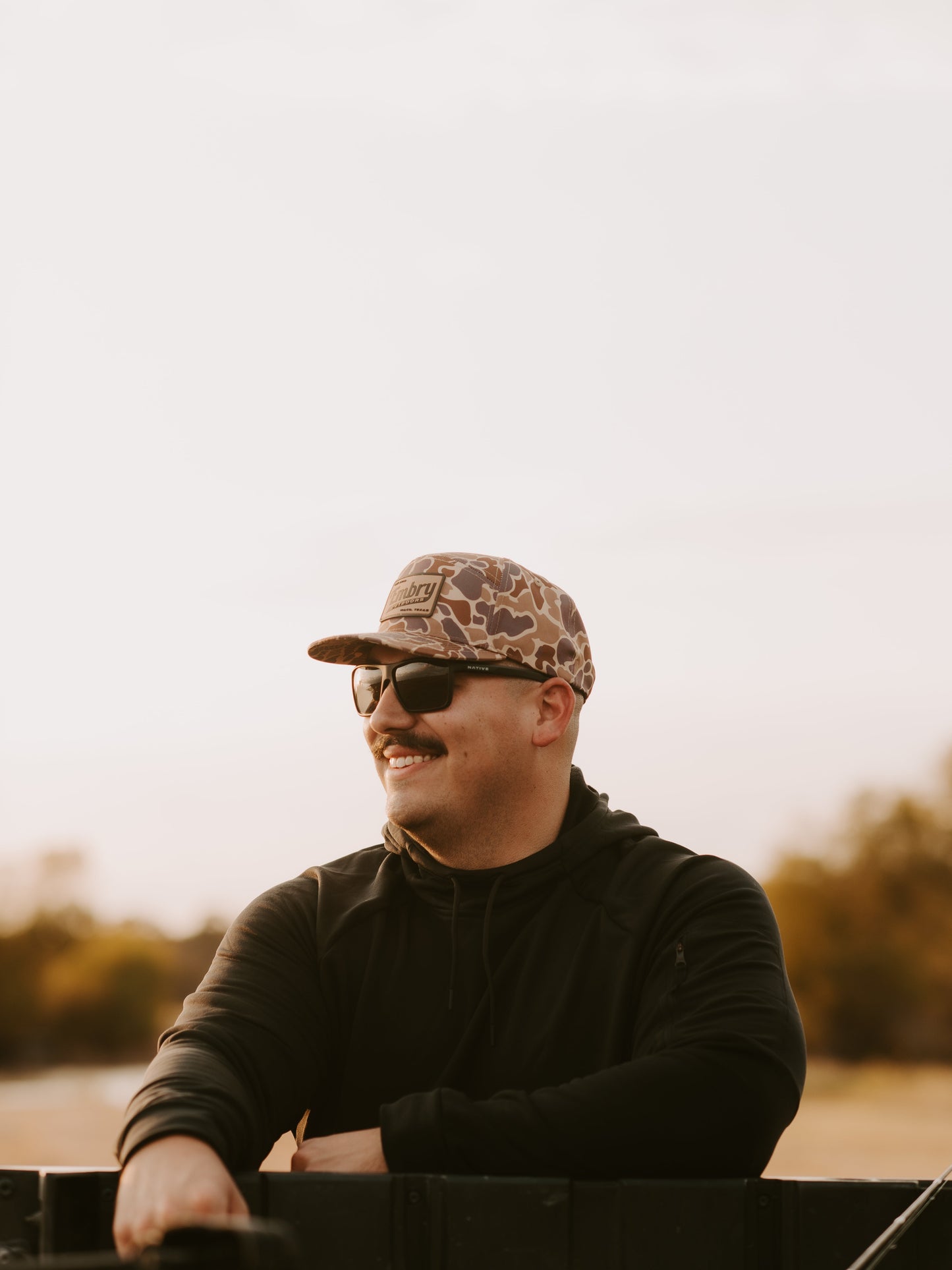 Man wearing Duck Camo 6 Panel "Retro" Trucker hat outdoors with sunglasses, showcasing stylish camo pattern.