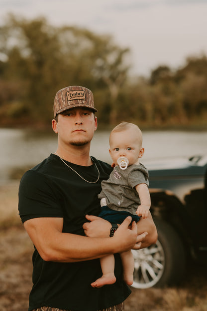 Man wearing Brown Ranch Camo Trucker Hat holding a baby outdoors near a river.