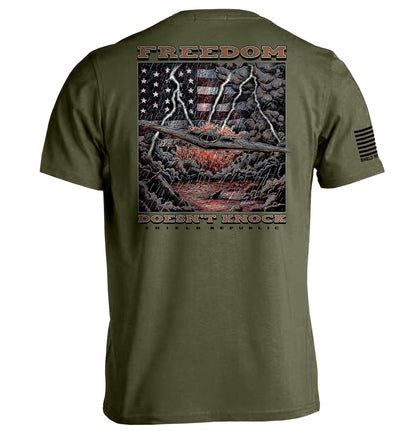 Freedom Doesn’t Knock Shirt with patriotic graphic, showing an American flag against a stormy backdrop on an olive tee.