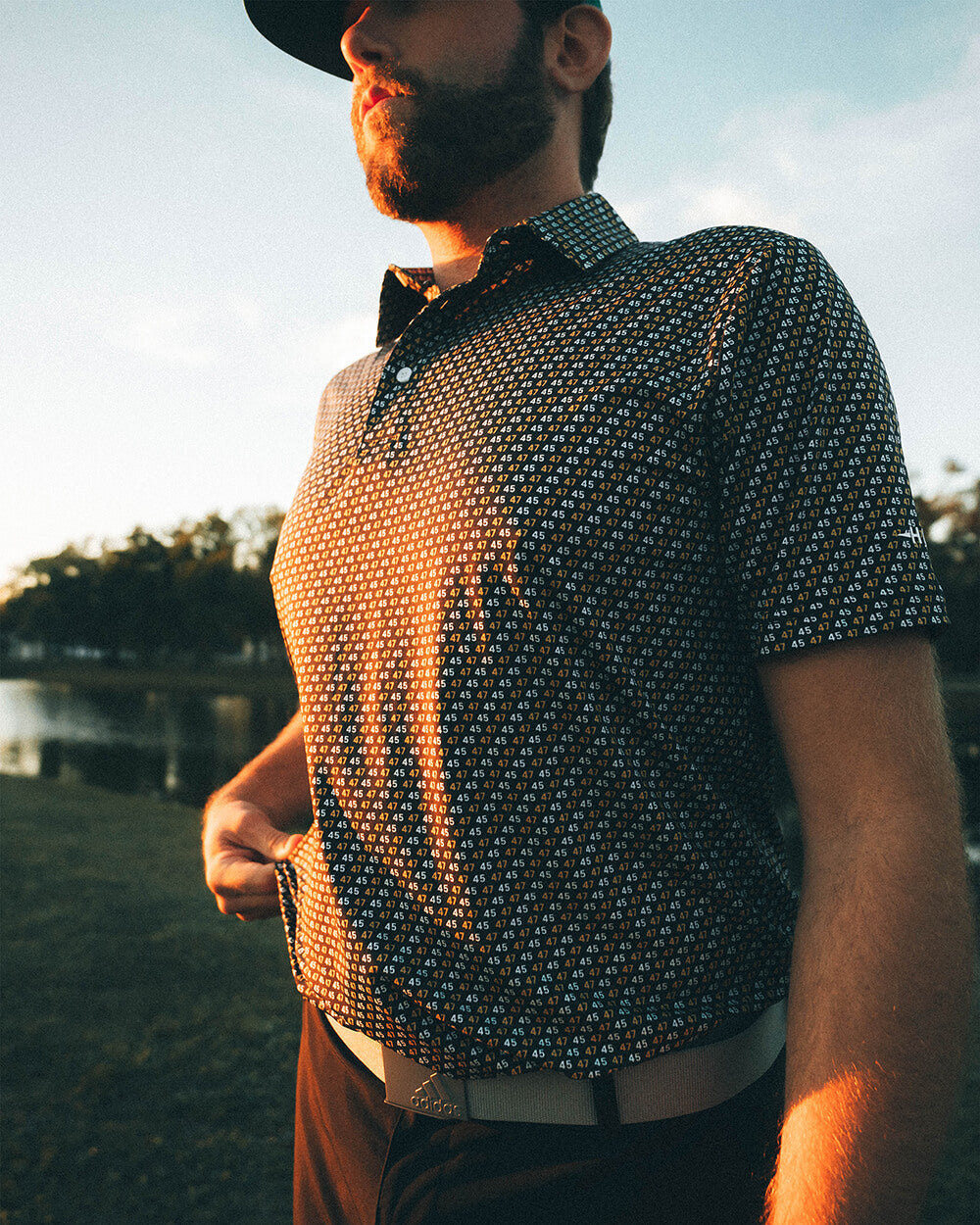 Man wearing 45 47 polo shirt in black-and-gold print by Haven Golf, standing outdoors with sunset lighting.